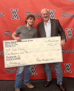 Two men smile, holding a large scholarship check from Parker Alexander Attorneys at Law against a "Rebels" background. Text on check: Parker Alexander Attorneys at Law, 2503 Ferndale St Monroe, LA 71201 (318) 322-7373. May 2, 2023. Pay to the Order of Leo