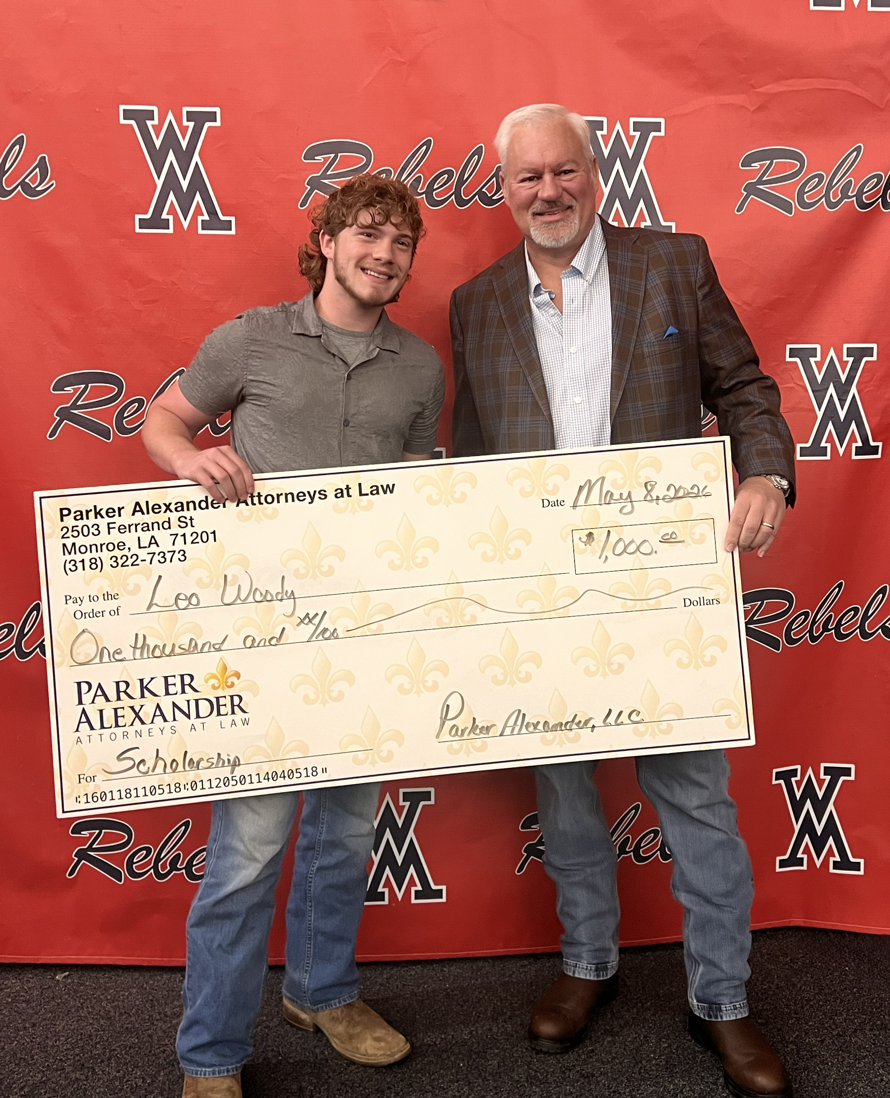 Two men smile, holding a large scholarship check from Parker Alexander Attorneys at Law against a "Rebels" background. Text on check: Parker Alexander Attorneys at Law, 2503 Ferndale St Monroe, LA 71201 (318) 322-7373. May 2, 2023. Pay to the Order of Leo